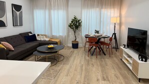 Moderni olohuone, jossa on sohva, sohvapöytä, ruokapöytä ja tuoleja. Huoneessa on myös televisio, lamppu ja ruukkukasvi.