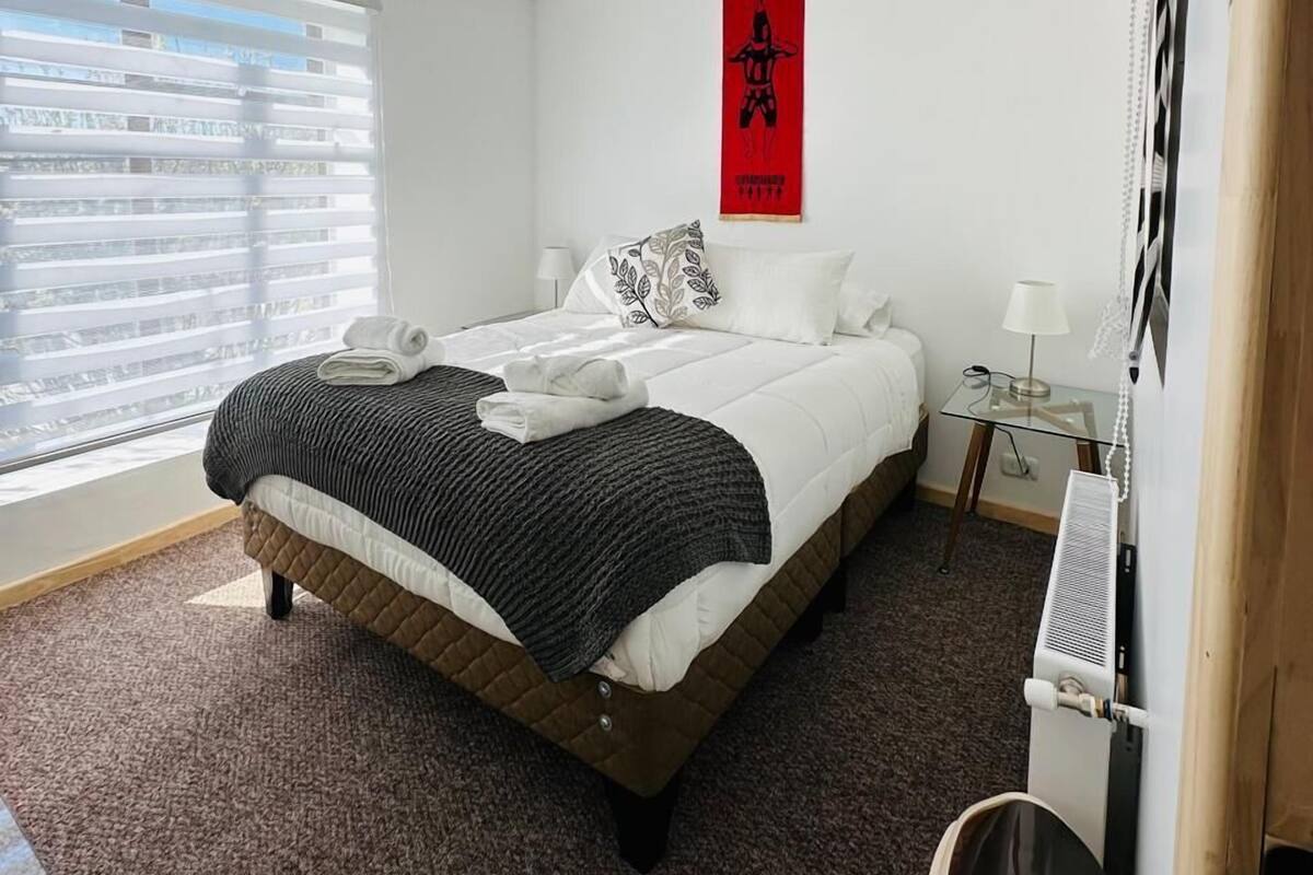 A modern hotel room with a large bed, a small bedside table, a red poster on the wall, and a window with blinds.