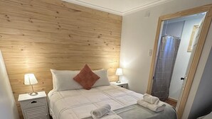 Habitación de hotel ordenada con cabecera de madera, dos mesitas de noche con lámparas, cama con colcha gris y almohadas blancas, y una puerta que da a una zona de ducha.