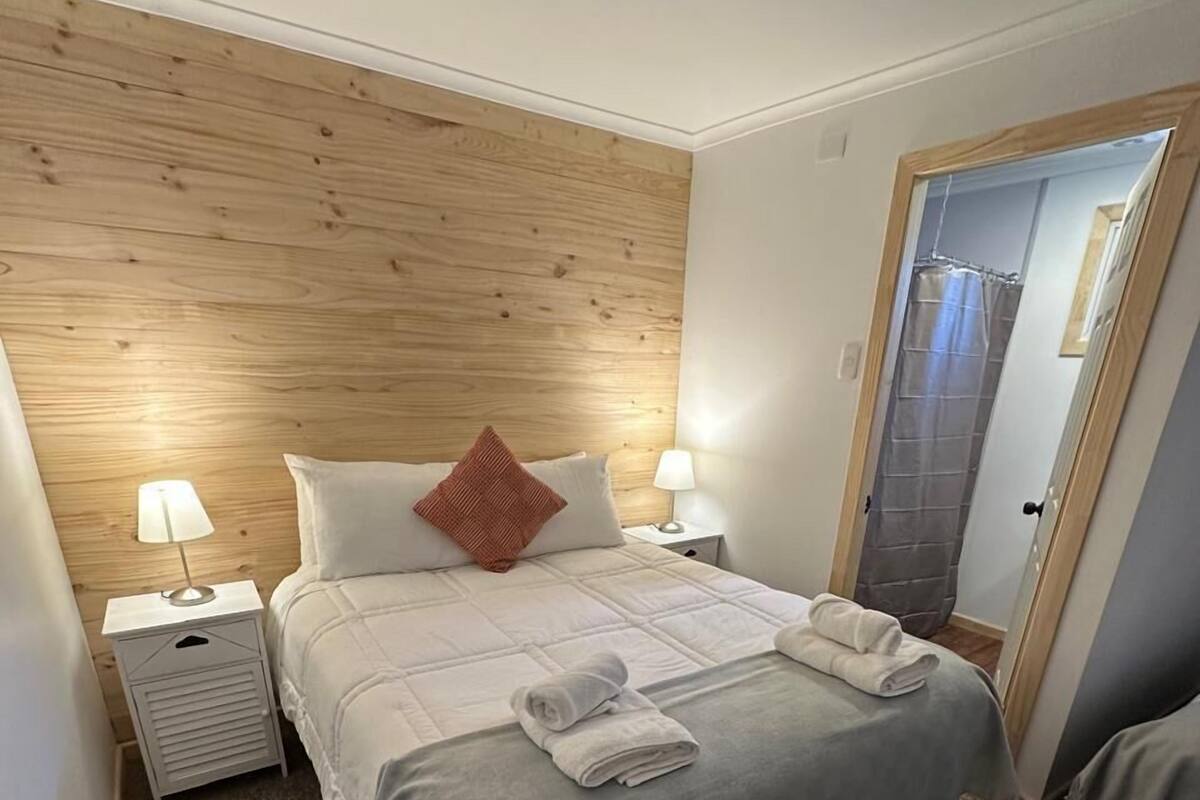A neatly arranged hotel room with a wooden headboard, two bedside tables with lamps, a bed with a grey bedspread and white pillows, and a door leading to a shower area.