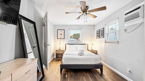 A bedroom with a bed, bedside tables, a ceiling fan, a window with blinds, and a wall-mounted air conditioning unit.