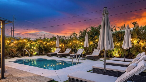 A swimming pool with lounge chairs and umbrellas, surrounded by palm trees and a sunset sky.