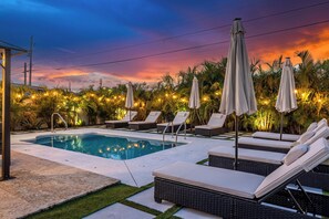 Una piscina con sillones de playa y sombrillas, rodeada de palmeras y un cielo al atardecer.