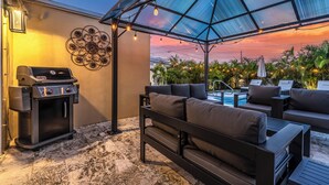 A patio area with a grill, outdoor seating, and a view of the pool and greenery.