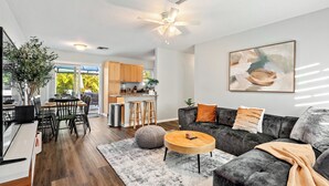 A modern living room with a dark grey sofa, a round wooden coffee table, and a grey shag rug. There is a dining area with a wooden table and chairs, and a kitchen in the background.