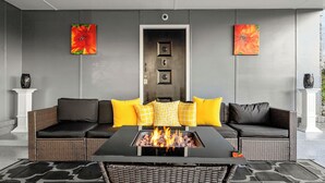 Ein moderner Außenbereich mit Sitzgelegenheit, einem Feuerbereich, zwei Sofas mit gelb-schwarzen Kissen und zwei an der Wand befestigten Kunstwerken.