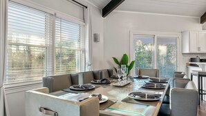 Ein Essbereich mit einem langen Tisch, gedeckt für sechs Personen, umgeben von gepolsterten Stühlen, einer Vase mit Grünpflanzen und Blick ins Freie durch große Fenster.