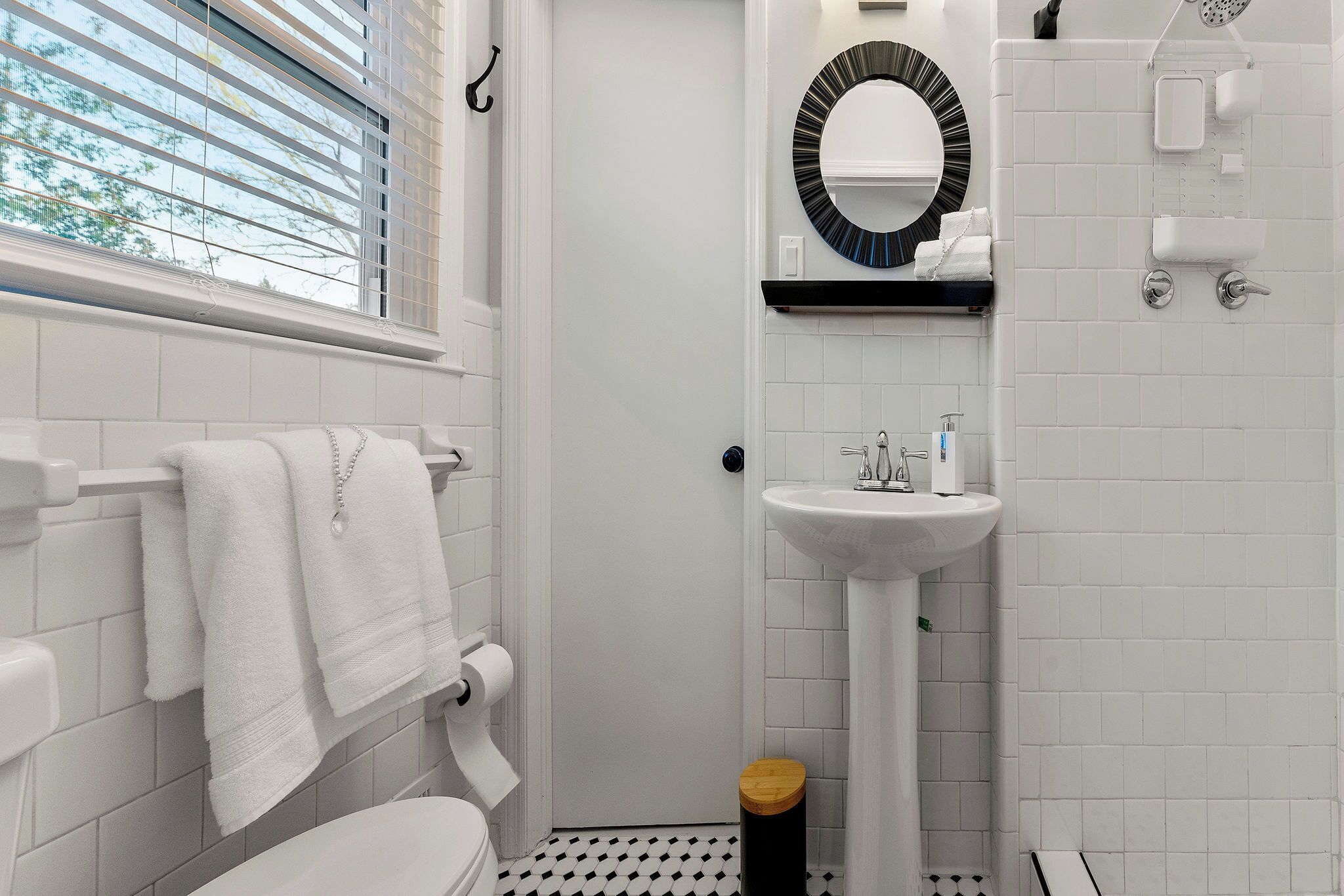 A bathroom with a toilet, sink, and shower area. There is a window with blinds, a towel rack, and a mirror with a circular frame.