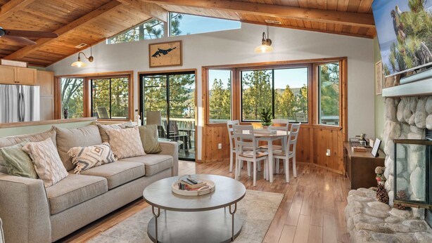 A spacious living room with a wooden ceiling, large windows, a stone fireplace, and a dining area. - Eagles Nest•Walk to Village (Big Bear Lake)