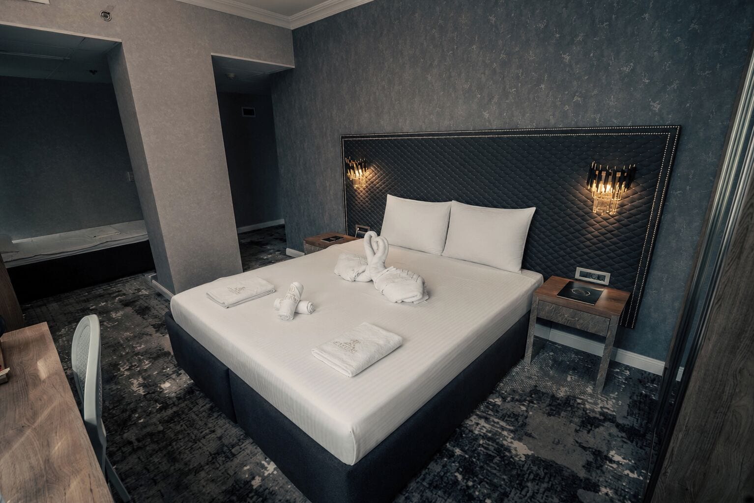 Una habitación de hotel moderna con una cama grande, cabecera con iluminación integrada, mesita de noche con radio y alfombra de diseño oscuro.