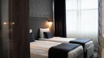 Habitación de hotel con una cama grande, otra más pequeña, cabecera con lámpara, ventana con cortinas y suelo de madera.