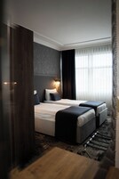 Habitación de hotel con una cama grande, otra más pequeña, cabecera con lámpara, ventana con cortinas y suelo de madera.