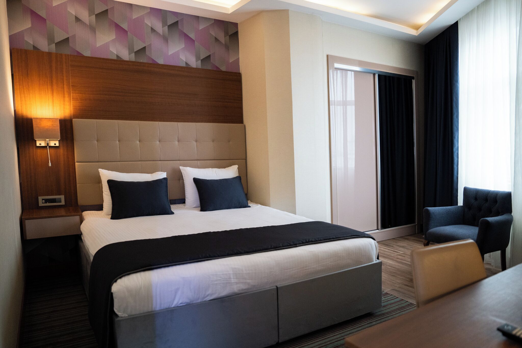 A hotel room with a large bed, a nightstand with a lamp, a chair, and a desk.
