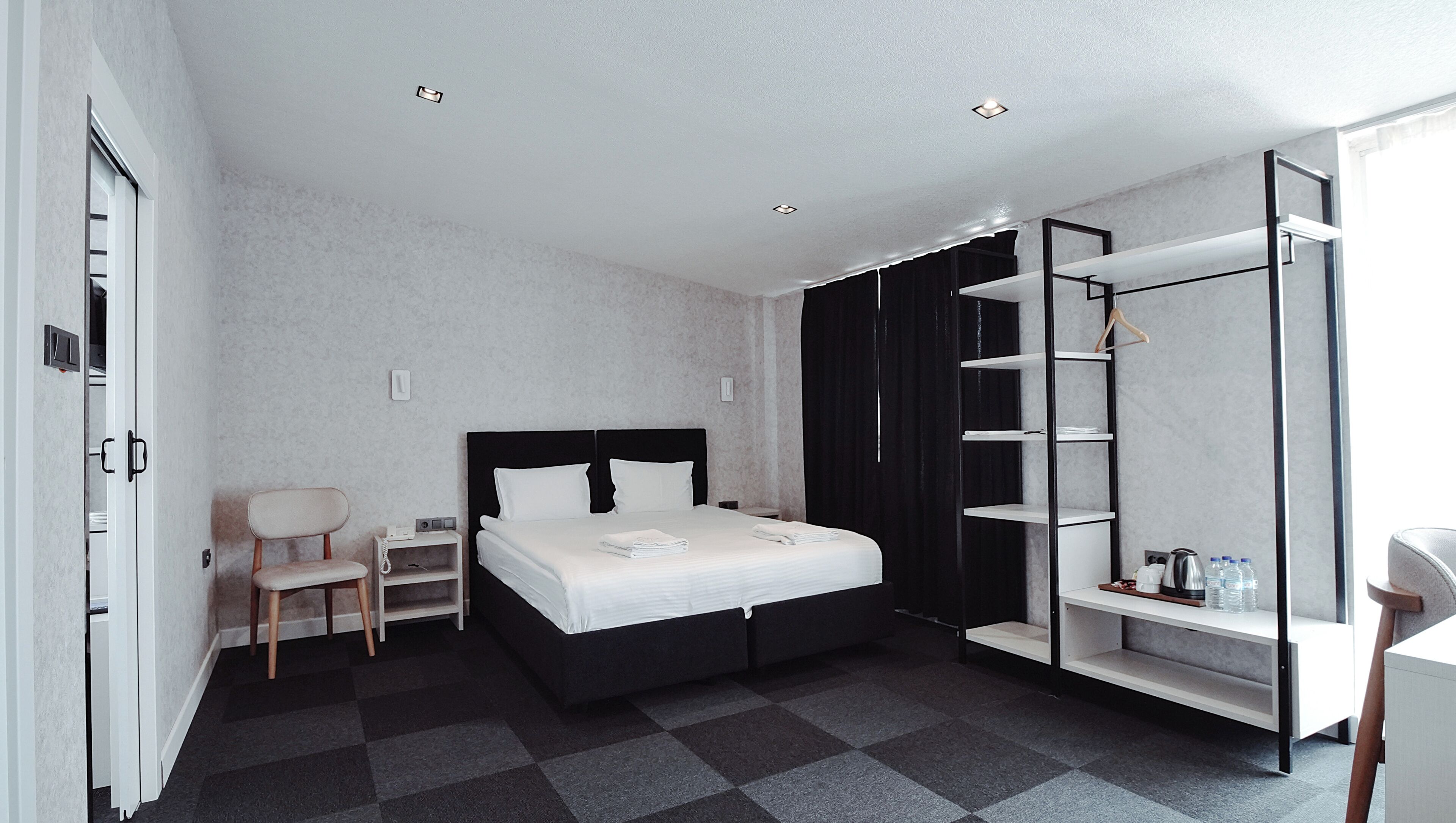 Una habitación de hotel moderna con una cama, una silla, un escritorio, un armario y una estantería con artículos de aseo.