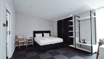 A modern hotel room with a bed, a chair, a desk, a wardrobe, and a shelf with toiletries.