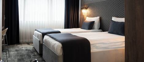 Une chambre d’hôtel moderne avec deux lits, une grande fenêtre avec des rideaux légers, une tête de lit sombre à texture, et un petit coin salon avec une chaise et une table.