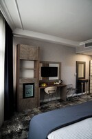 A hotel room with a bed, a desk with a chair, a television, a mirror, and a small kitchenette area.