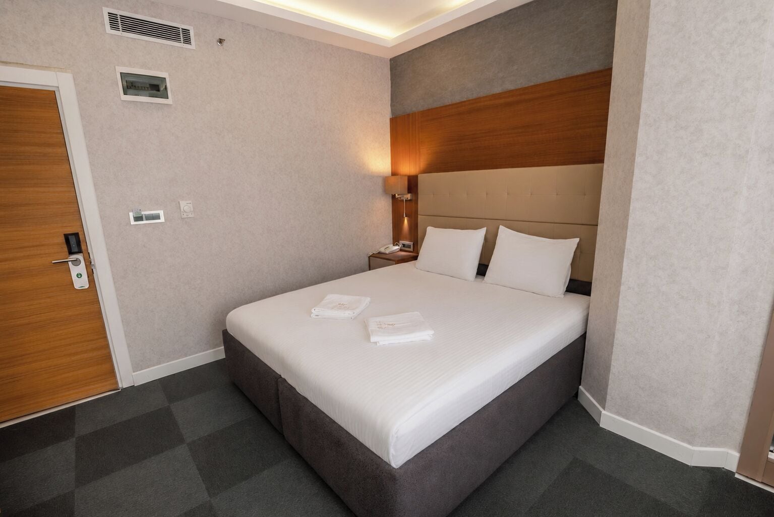Habitación de hotel con cama, mesita de noche, calefactor en la pared y una puerta con manija.