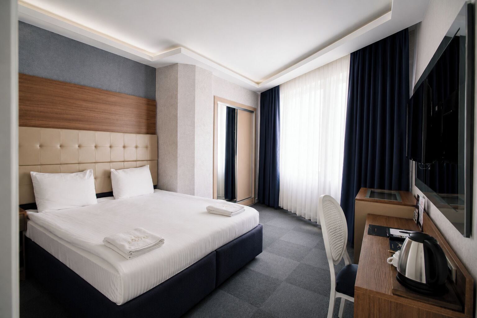 A hotel room with a large bed, a desk with a chair, a television, and a window with curtains.