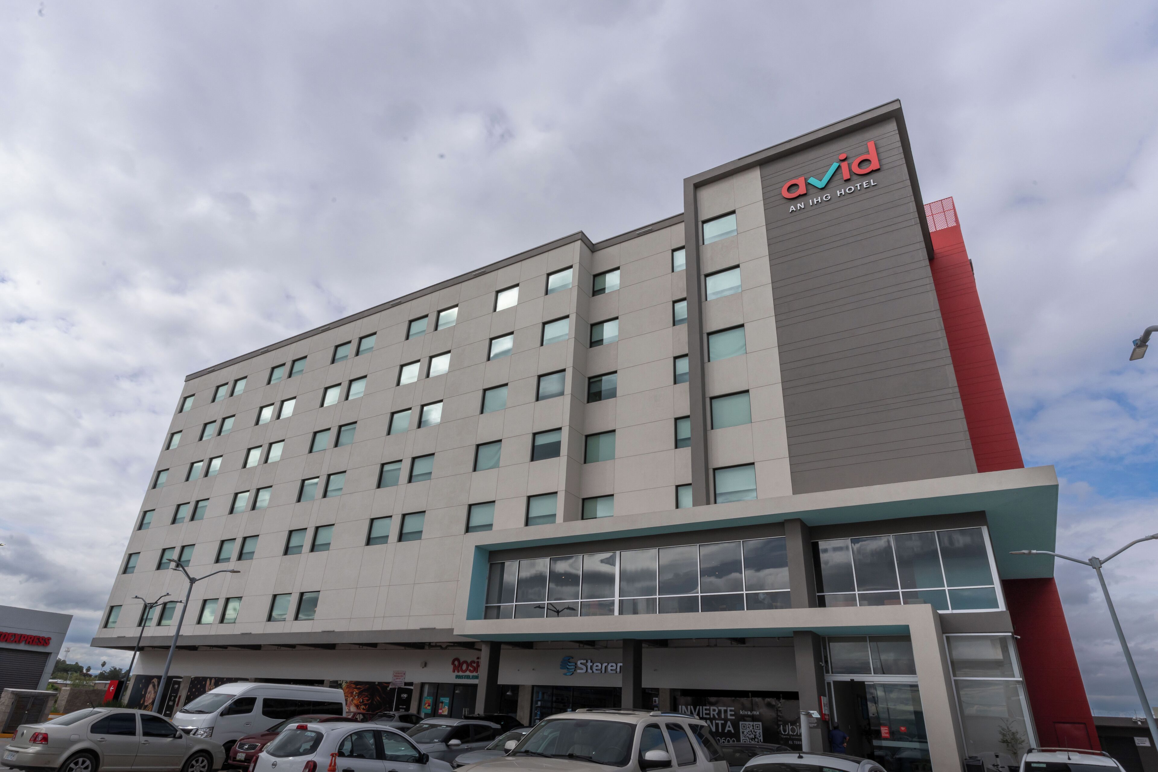 A modern hotel building with multiple floors, a prominent red and grey facade, and several ground-level retail spaces including a pharmacy and a bank.