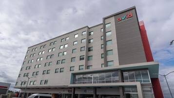 Un moderno edificio de hotel con varios pisos, una fachada prominente en rojo y gris, y varios locales comerciales a nivel de calle, incluyendo una farmacia y un banco.