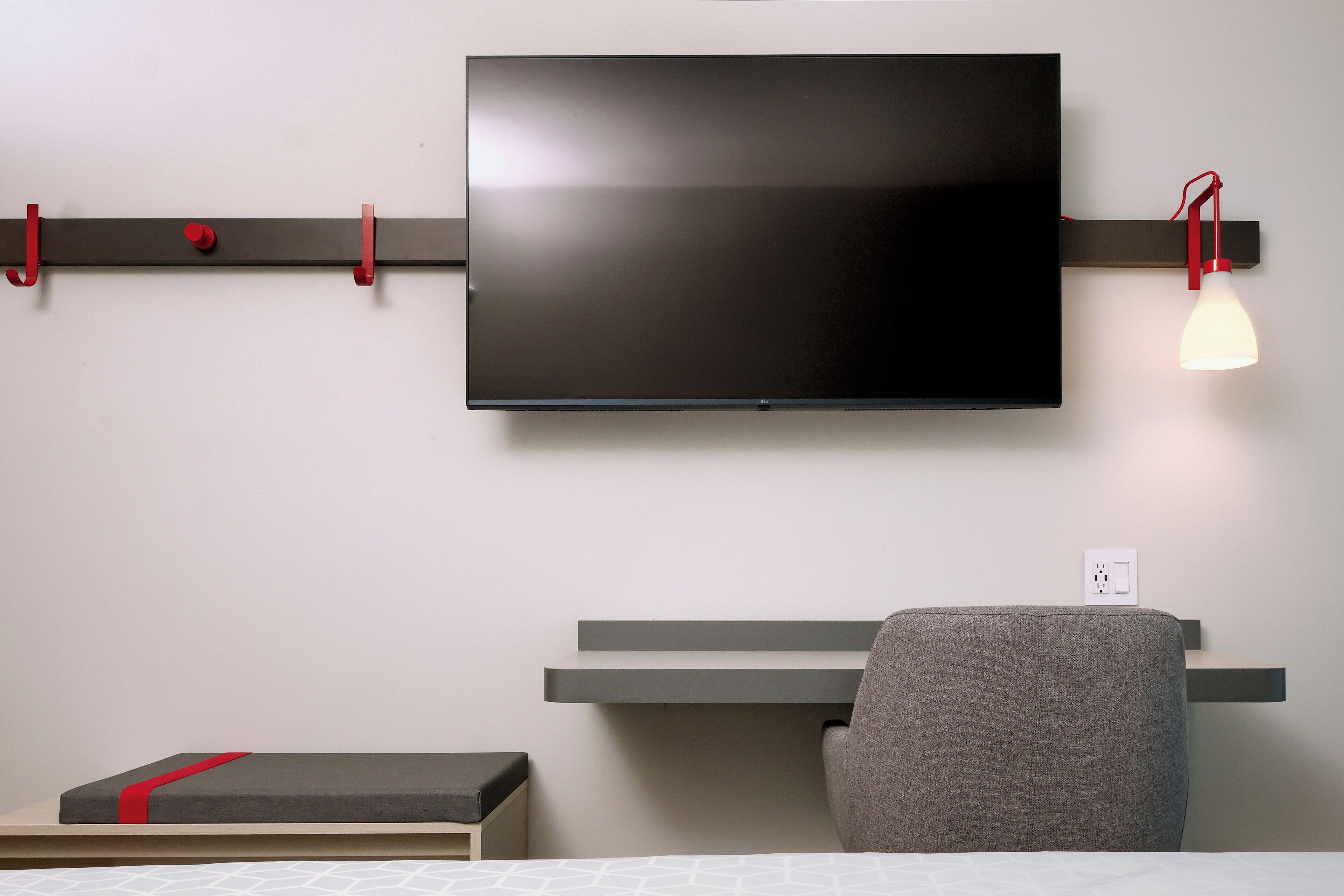 A modern room with a flat-screen TV mounted on the wall, a desk with a chair, and a bedside table with a red accent.