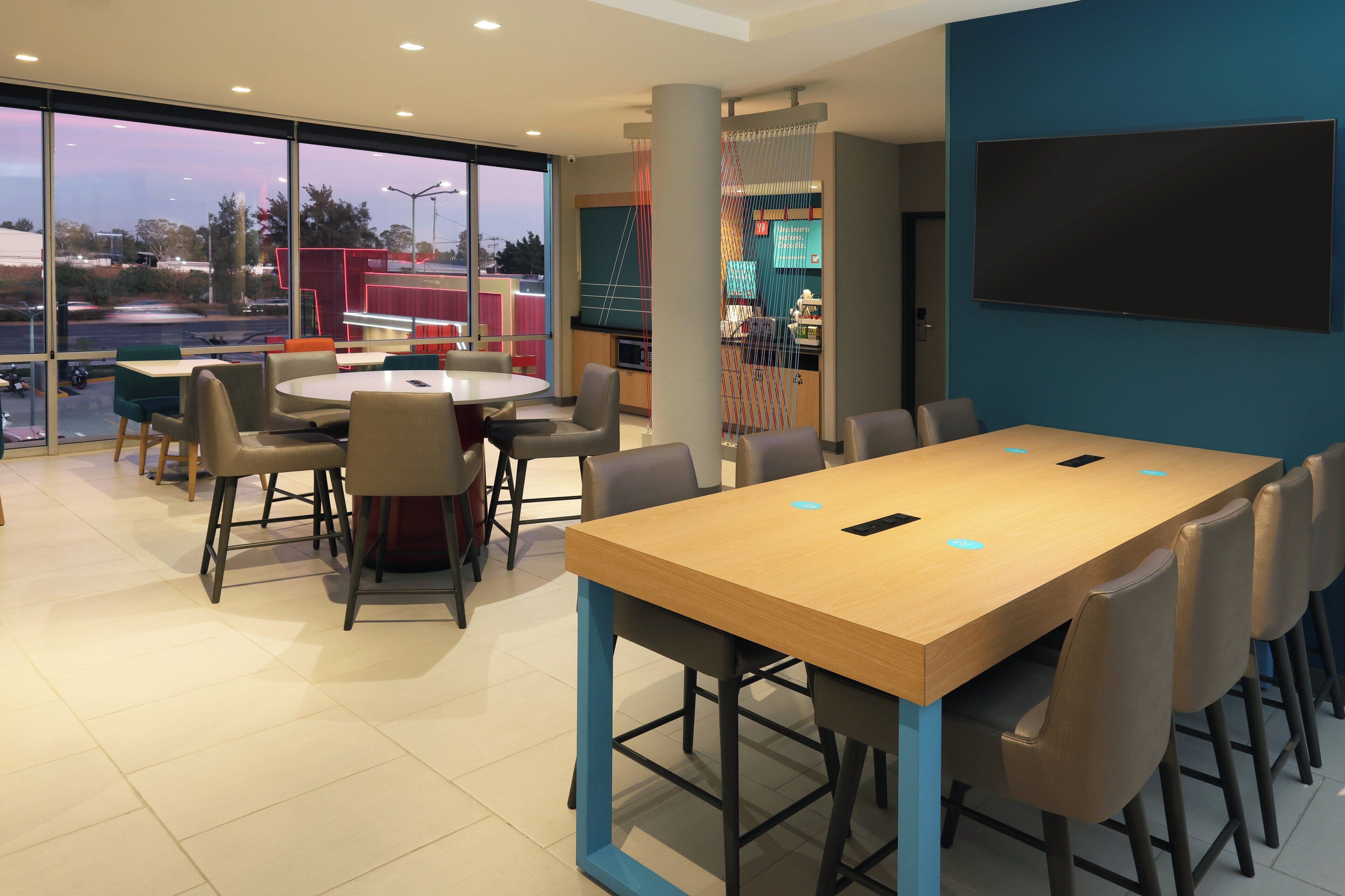 A modern conference room with a large table and chairs, large windows with a view of the outside, and a flat-screen TV mounted on the wall.