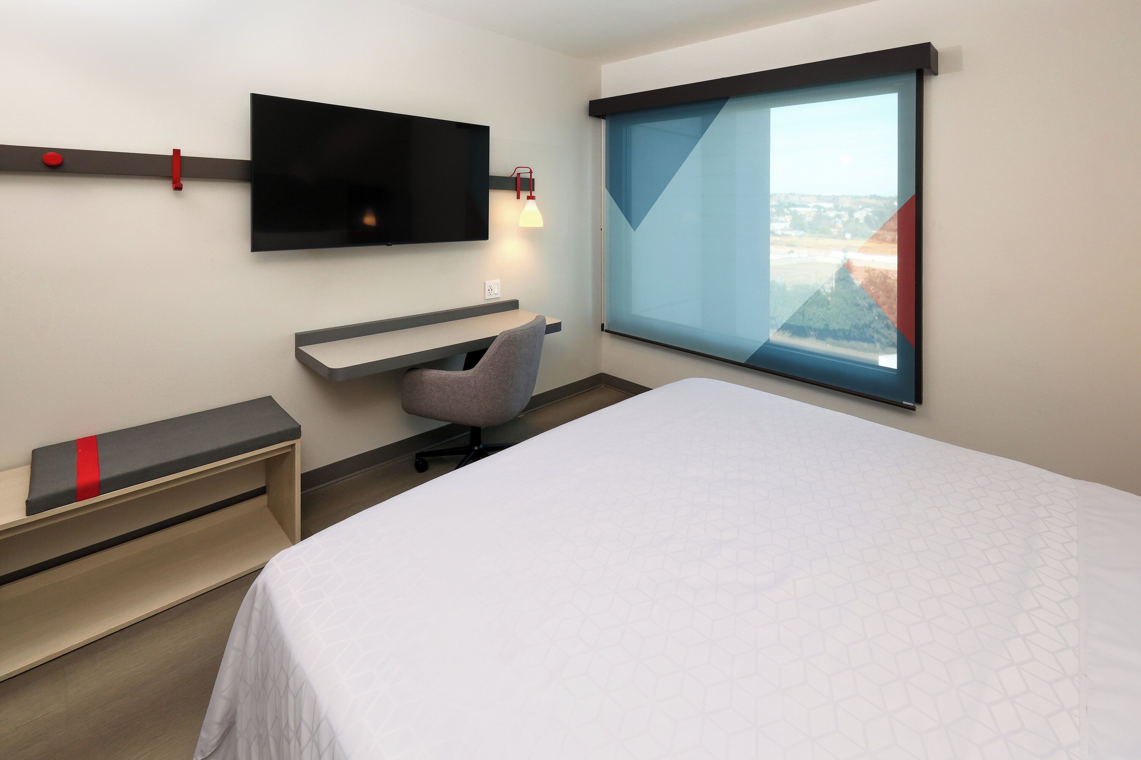 A modern hotel room with a bed, a desk, a chair, a TV, and a window with a city view.
