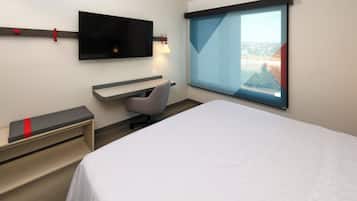 A modern hotel room with a bed, a desk, a chair, a TV, and a window with a city view.