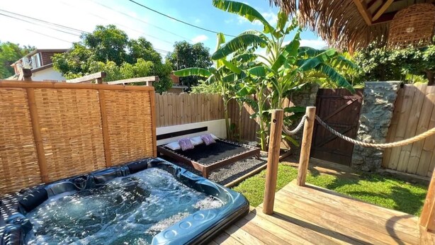 Un bain à remous sur une terrasse en bois, un petit lit avec des oreillers et une barrière faite de cordes, le tout dans un jardin.