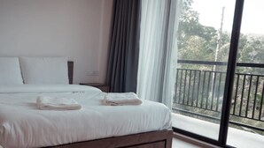 A neatly made bed with white linens and two folded towels, a nightstand, and a large window with a view of trees and a balcony. - Casa Wyna family Ayur center (Mananthavady)