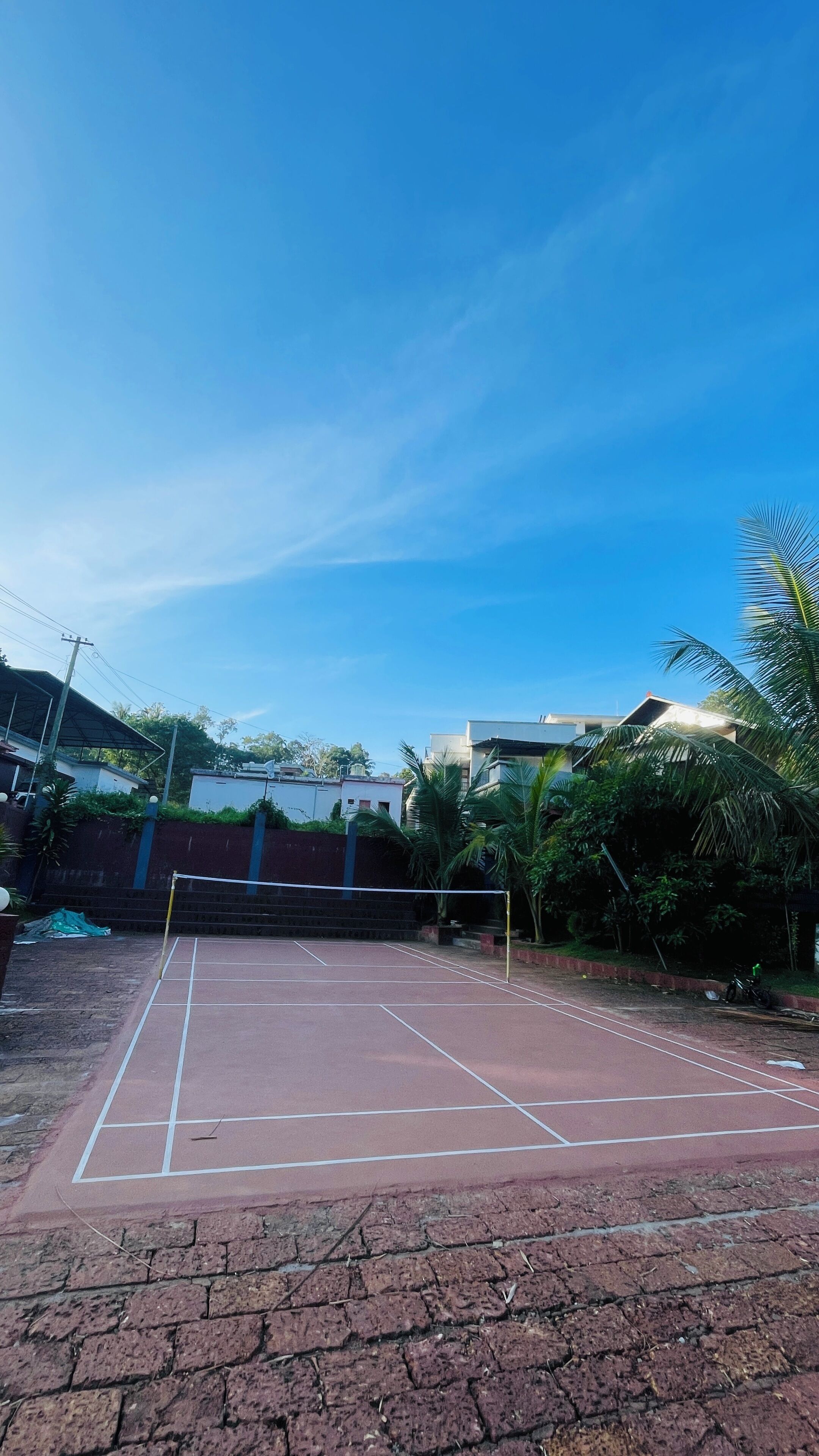 Uma quadra de tênis com rede, cercada por um muro de tijolos e vegetação.