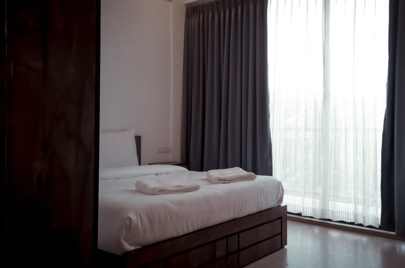 Una cama bien hecha con sábanas blancas y dos toallas dobladas, una mesita de noche con una lámpara, y un ventanal con cortinas grises.