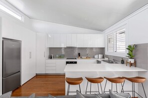 Una cocina moderna con armarios blancos, un refrigerador de acero inoxidable, una isla central con taburetes y una ventana con persianas.