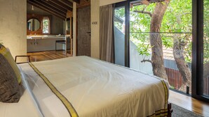 Un dormitorio con una cama grande, un baño con espejo y un ventanal corredero que da a un balcón con árboles.