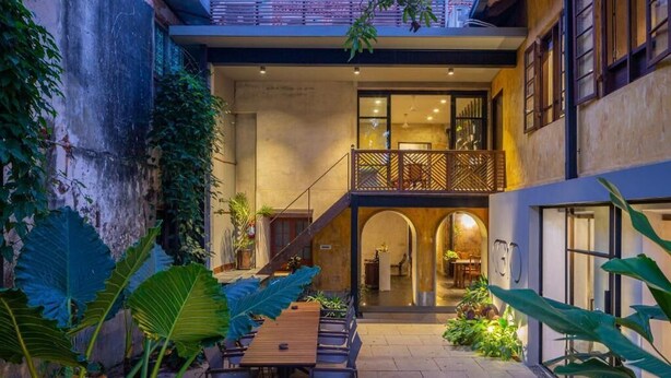 Un edificio moderno con un patio cubierto que tiene una mesa y sillas de madera, rodeado de plantas verdes grandes.