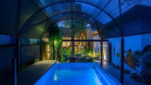 Una piscina moderna con cerramiento de vidrio, rodeada de exuberante vegetación y vista a una casa.