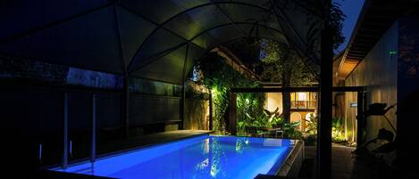 An outdoor swimming pool with a glass ceiling, surrounded by greenery and a modern building.