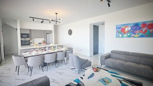 A modern living room with a grey sofa, a dining area with a white table and grey chairs, a kitchen with grey cabinets, and a colorful abstract painting on the wall. - Protaras Aqua Pearl Villa AQ12 (Paralimni)