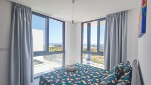 A modern bedroom with large windows, a bed with a patterned bedspread, and a view of the city. - Protaras Aqua Pearl Villa AQ12 (Paralimni)