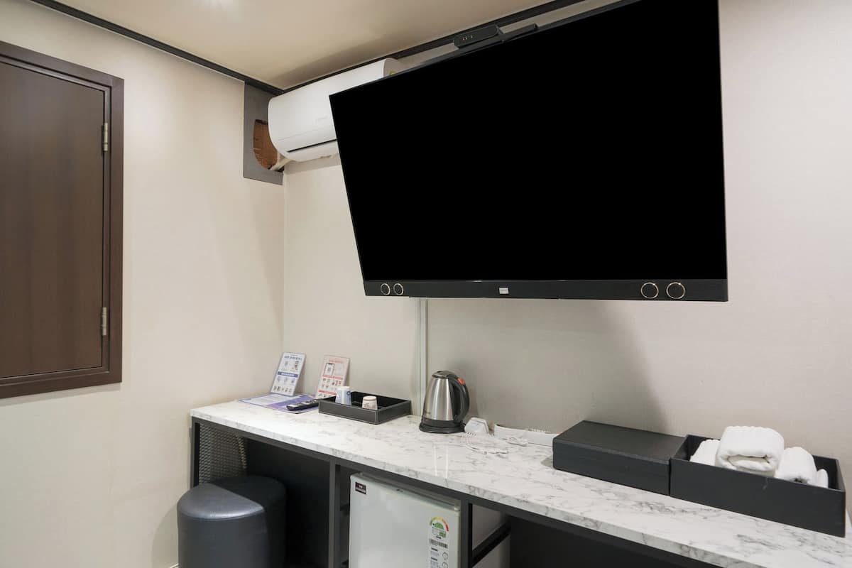 A hotel room with a flat-screen TV mounted on the wall, a small desk with a kettle, a trash bin, and a mini-fridge.