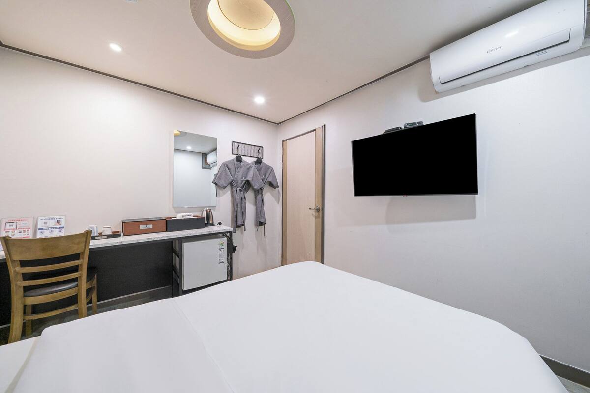 A hotel room with a bed, a desk with a computer, a chair, a mirror, a TV mounted on the wall, and an air conditioner.