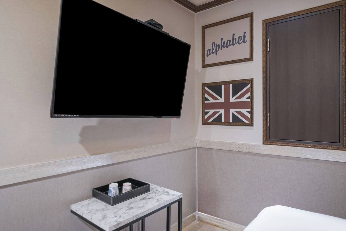 A hotel room with a flat-screen TV mounted on the wall, a small table with a tray, and two framed pictures, one with the word 'alphabet' and another with a Union Jack flag.