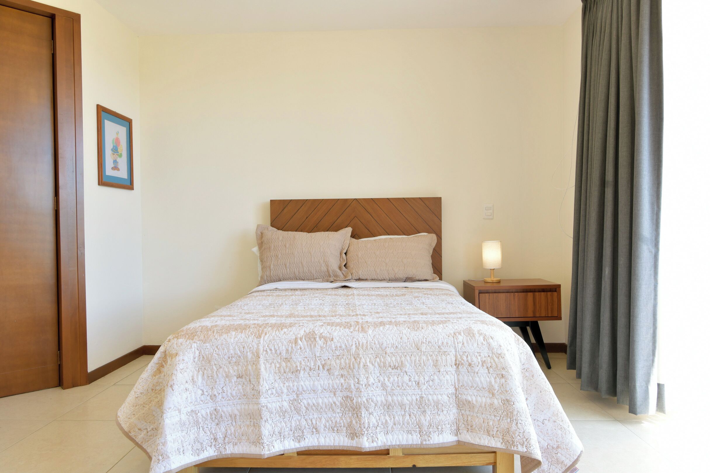 A neatly made bed with a wooden headboard, a bedside table with a lamp, and a grey curtain.