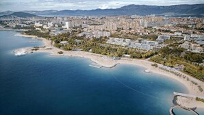 Vue aérienne d’une ville côtière, avec une plage, un centre urbain dense et des montagnes en arrière-plan.