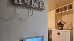 Seinälle asennettu televisio, koristeellinen ”KOTI”-kyltti, valokytkin ja valkoinen jääkaappi keittiötilassa.