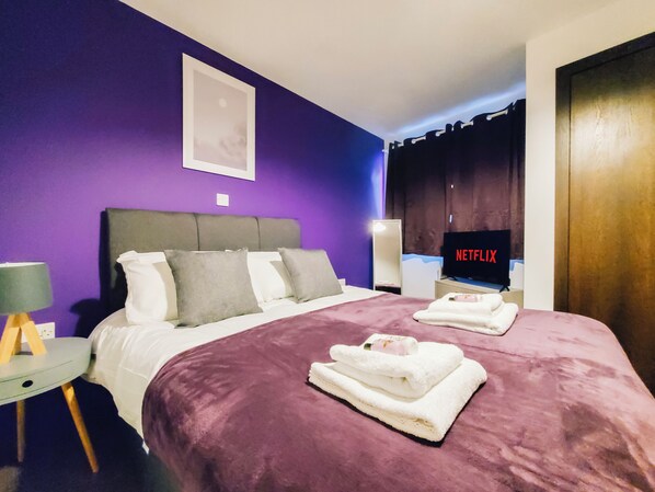Relax in a spacious room with a large bed, flat-screen TV, and a small table with a lamp.