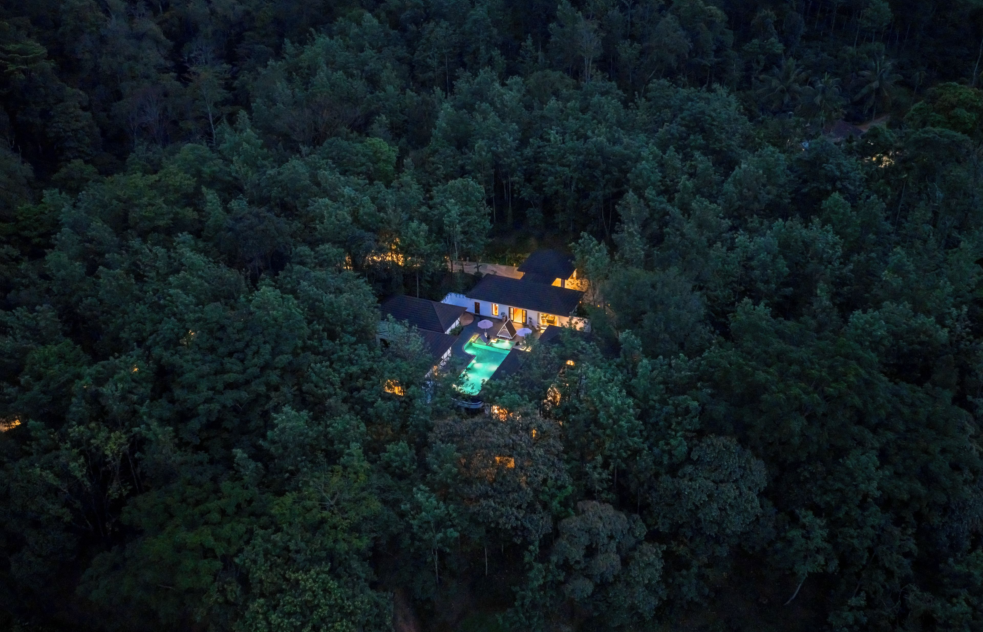 An aerial view of a secluded house surrounded by dense forest, with a swimming pool and a well-lit patio area.