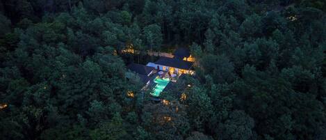 An aerial view of a secluded house surrounded by dense forest, with a swimming pool and a well-lit patio area.