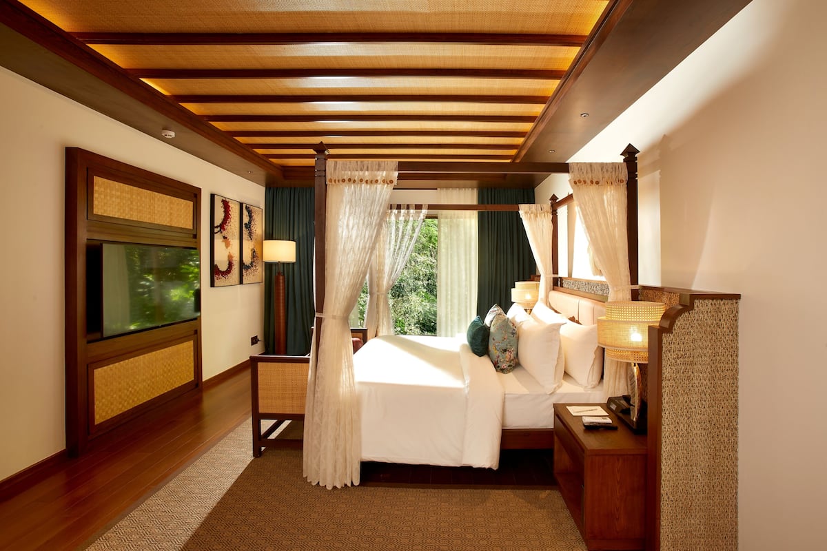 Habitación de hotel con cama, mesita de noche, lámpara y vistas a zonas verdes por la ventana.